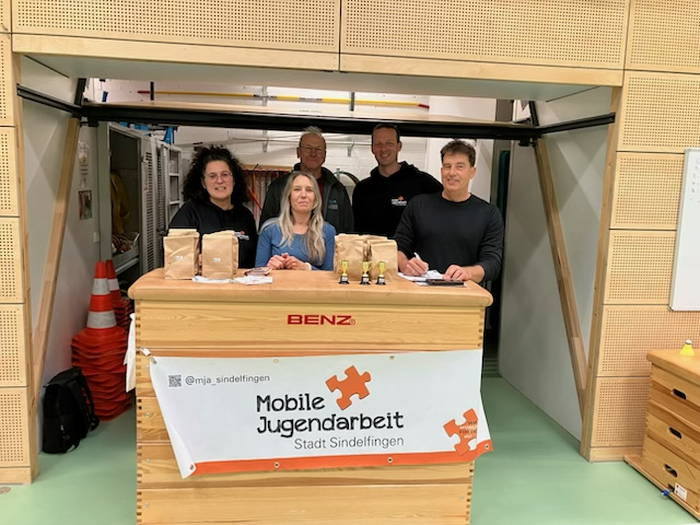 Eine Gruppe von Schulsozialarbeit und mobiler Jugendarbeit in der Sporthalle hinter einer Theke aus eine Sportkasten