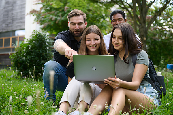 Studierende auf einer Wiese vor dem HHZ mit Laptop