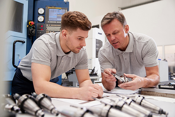 Junger Mann bei einem technischen Betrieb, schaut sich ein Werkstück mit dem Ausbilder zusammen an